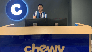 Andrew Han stands behind a large desk with the blue "Chewy" logo on it.