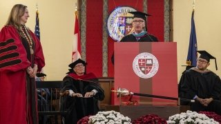 Saint Lawrence University President Kathryn Morris, wearing scarlet academic regalia, listens as Michael Ranger gives a speech.