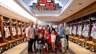 Joe Marsh stands with his family in the Saints Men's Ice Hockey locker room. The room has white and scarlet hockey jerseys on each side. A neon Saint Lawrence shield hands from the ceiling.