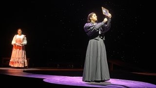 An actress, wearing a grey gown, stands in the spotlight on stage holding a piece of paper up to the light. Another actress, wearing a pale orange gown, stands in the background.