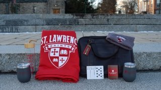 A photo on the steps from the top of the quad with two gray St. Lawrence tumblers, a large red blanket with the St. Lawrence Saints logo with the crest in white, a leather bag, a St. Lawrence deck of cards with a royal flush in spades revealed and a gray tuc with the St. Lawrence Saints logo.