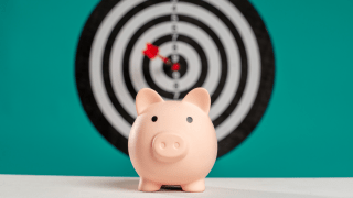 A pink piggy bank is front and center. In the background, a bullseye target is mounted on a blue-green wall, with a dart in the center of the bullseye.