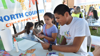 A St. Lawrence student works on a craft with a child on a summer day under a large tent. In the background, a banner for the North Country Children's Museum is visible