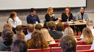 Five panelists sit at a table, facing an audience of students in theatre-style seating. The panelist in the center is speaking with a microphone in one hand, with the other hand is held out.
