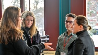 Four people stand indoors as a group, directing their attention to the speaker.