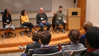 Four discussion panelists seated on stage face an audience of students. One of the speakers on stage is speaking to the group.