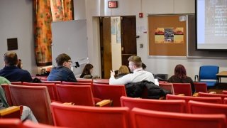 Students are seated in theatre chairs inside a lecture hall. Students appear to be in small discussion groups, speaking face to face.