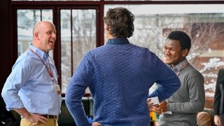 Three people stand together indoors. Two of the people are facing the speaker of the group, smiling as they direct their attention to the speaker.