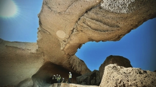 A large rock structure appears in the foreground and students wearing neon yellow construction vests and helmets stand beneath the rock structure.