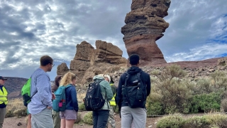 On a cloudy day, a group of students stand together as they observe a large, vertical geological structure.