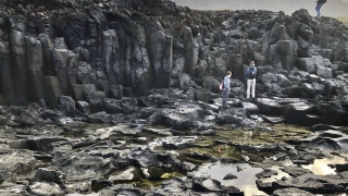 Three students stand on staggered rock structures. The rocks are dark gray, scattered in various shapes and sizes. In the background, a mountain structure appears through the hazy clouds.