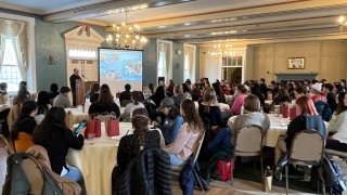 A person stands at a podium and addresses a room full of students sitting at round tables with white table clothes.