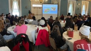 Several Saint Lawrence students sit at round tables, covered in white tableclothes, and listen as a faculty member stands at a podium and gives a presentation on a projector screen.
