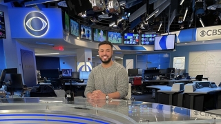 Michael Paulino, wearing a gray sweater, sits at the CBS News Desk in the main newsroom. He's surrounded by blue and white screens. There are two digital CBS logos in the background.