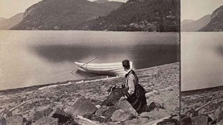 A black and white photo of a woman wearing a long sleeve dress and sitting on a rocky shoreline, gazing out at the Twin Mountains on Sabbath Day Point. A white boat with ores sits in the water. 