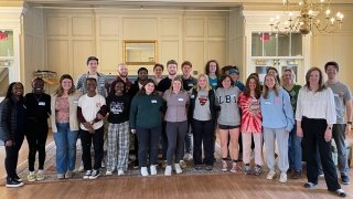 A group of Saint Lawrence students gather together with President Morris in celebration of academic excellence. There is a large gold chandelier hanging from the ceiling of the white room.
