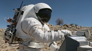 Dean Epler, wearing a NASA space suit, works on equipment in the desert. 