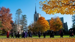 Several people wearing graduation regalia, walk in a line on a bright fall day. There are piles of orange and yellow leaves on the ground. Gunnison Memorial Chapel is in the background.