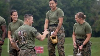 Three women dressed in camouflage pants and a green T-shirt stand on a podium, holding kettle bell weights. The one in the center reaches her hand out to shake the hand of a man dressed in the same outfit.