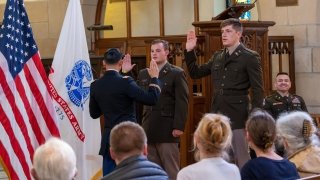 Two Reserve Officers Training Corps and soon to be graduates wear military uniforms and raise their right hands during a swearing in ceremony. An officer sits in the background and smiles with pride as the 2nd lieutenant's families look on.