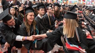 Two students, wearing commencement regalia, reach across the aisle and hold hands. The person in between laughs as they all celebrate Commencement 2023.