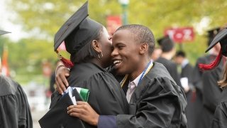 Two students, wearing academic regalia, embrace outside as they await Commencement.