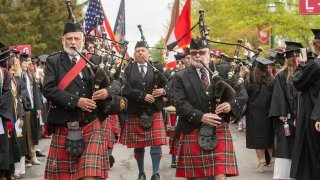 A group of bag pipers place music outside while a sea of graduates line the side walk on each side of the musicians.