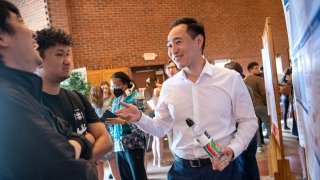 A student stands in front of a research poster and laughs with two Festival Day attendees as they explain their research. People circulate and examine posters in the background.