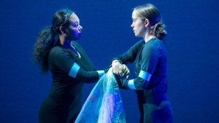 Two dancers, wearing black long-sleeve shirts and pants, clasp hands on a blue-lit theatre stage.
