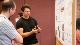 A student stands in front of their research poster and holds a small black box with wire protruding from it and explains their research to an attentive Festival Day participant.