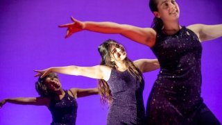 Three dancers, wearing purple sequin leotards, hold their arms out and smile on a purple lit stage.