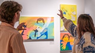 In an art gallery, people hold their hands out in front of their faces while they discuss three pieces of artwork painted in yellow, coral, and blue. 