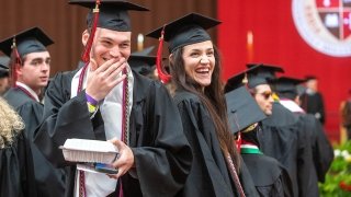 A student, wearing commencement regalia, holds their hand in front of their mouth to cover laughter as they hold a fast food container and await the Commencement festivities.