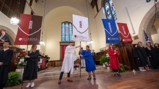 Students stand at the front of Gunnison Memorial Chapel in black, white, blue, and red robes, holding banners that represent their class years.