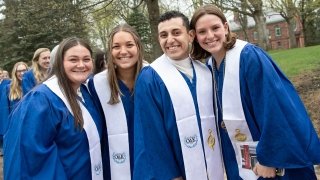 Four students, wearing royal blue Omicron Delta Kappa robes and white sashes stand together outside and smile. A line of people in royal blue robes are in the background.