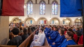 Students in black, white, blue, and red robes sit in pews in Gunnison Memorial Chapel. A choir performs at the front of the Chapel.