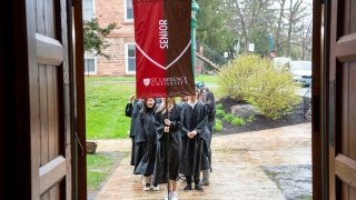 Students wearing black robes hold a scarlet and brown banner that represents their senior class standing and wait outside of Gunnison Memorial Chapel on a rainy day.