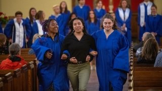 Two students wearing royal blue robes flank a student as they run down the center isle of Gunnison Memorial Chapel.