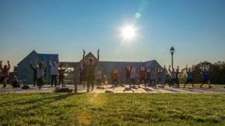 group of students doing yogo on the quad lawn platform, session led by one person. In the background we can see the Kirk Douglas Hall and a beautiful sunrise shining through, onto the students, the building and effecting the image slightly.