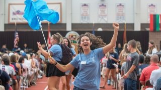 A student, wearing a robin blue t shirt, jumps in the air in excitement while waving a bright blue flag in the air. A sea of Saint Lawrence student in their families sit in chairs inside Newell Fieldhouse in the background.