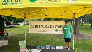 Ryan Krugman stands next to a sign that reads &quot;Debit, Credit, SNAP&quot; under a yellow tent, representing GardenShare.