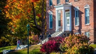 Students walks down the stairs of an early-Italianate style academic building made of red brick, surrounded by fall foliage.