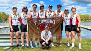 Members of the Saints Men's Rowing Team, wearing white and scarlet uniforms, wear medals around their necks and holding up a brown Saint Lawrence Saints flag, grin as they stand in front of a body of water and celebrate their victory.