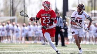Chris Jordan, wearing a scarlet lacrosse jersey, cradles the ball away from the competition on the lacrosse field. A large group of athletes, wearing white jerseys, stand out of focus in the background.