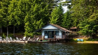 A robin blue boat house rests upon the Upper Saranac Lake shore. Several Adirondack style chairs are placed across the deck