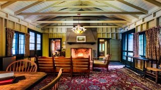 The living room of an Adirondack style cabin with wooden beams on the ceiling. A brick fire place has a warm, glowing fire while a several books rest on a table nearby.
