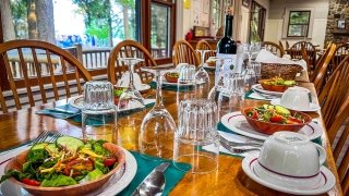 A long wooden dining table with fresh green garden salads. The table is set with clear drinking and wine glasses, and white coffee cups and plates. A bottle of red wine is also on the table.