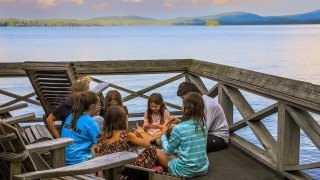 A group of children sit cross legged in a circle on a dock overlooking upper saranac lake.