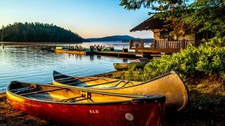 A red and a yellow canoe rest on the beach at Upper Saranac Lake. A large boat house and floating dock are in the background as the sunsets.