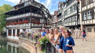 A group of Saint Lawrence students stand next to a railing overlooking water while abroad in France. White and brown building are in the background.